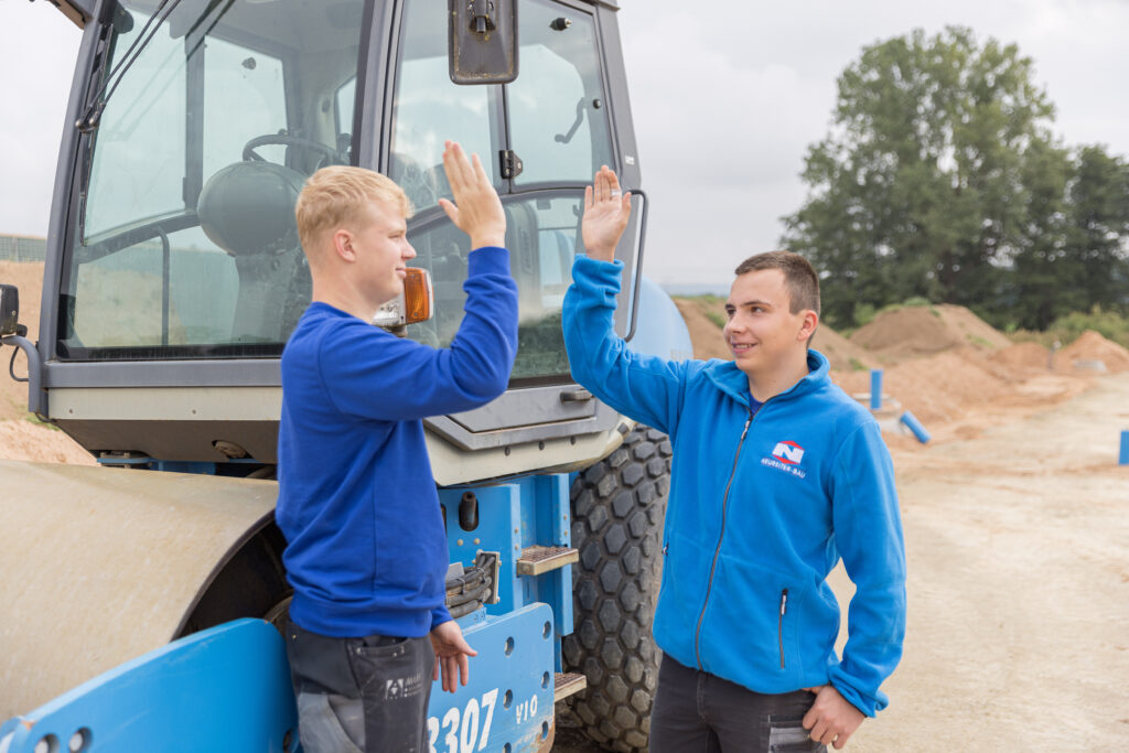 Neureiter Bau Ausbildung Karriere Baustelle Azubis neben Bagger die sich ein High-Five geben - Fremdingen Donau-Ries